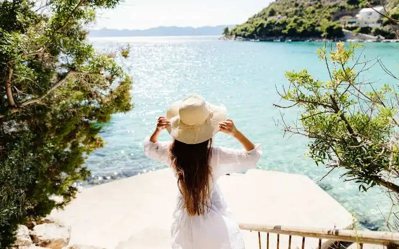 Figure in a white dress and sun hat overlooking a coastal view.